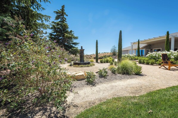 A landscaped garden with a fountain, tall cypress trees, and a building with a covered patio area. - La Maison du Vin (Paso Robles)