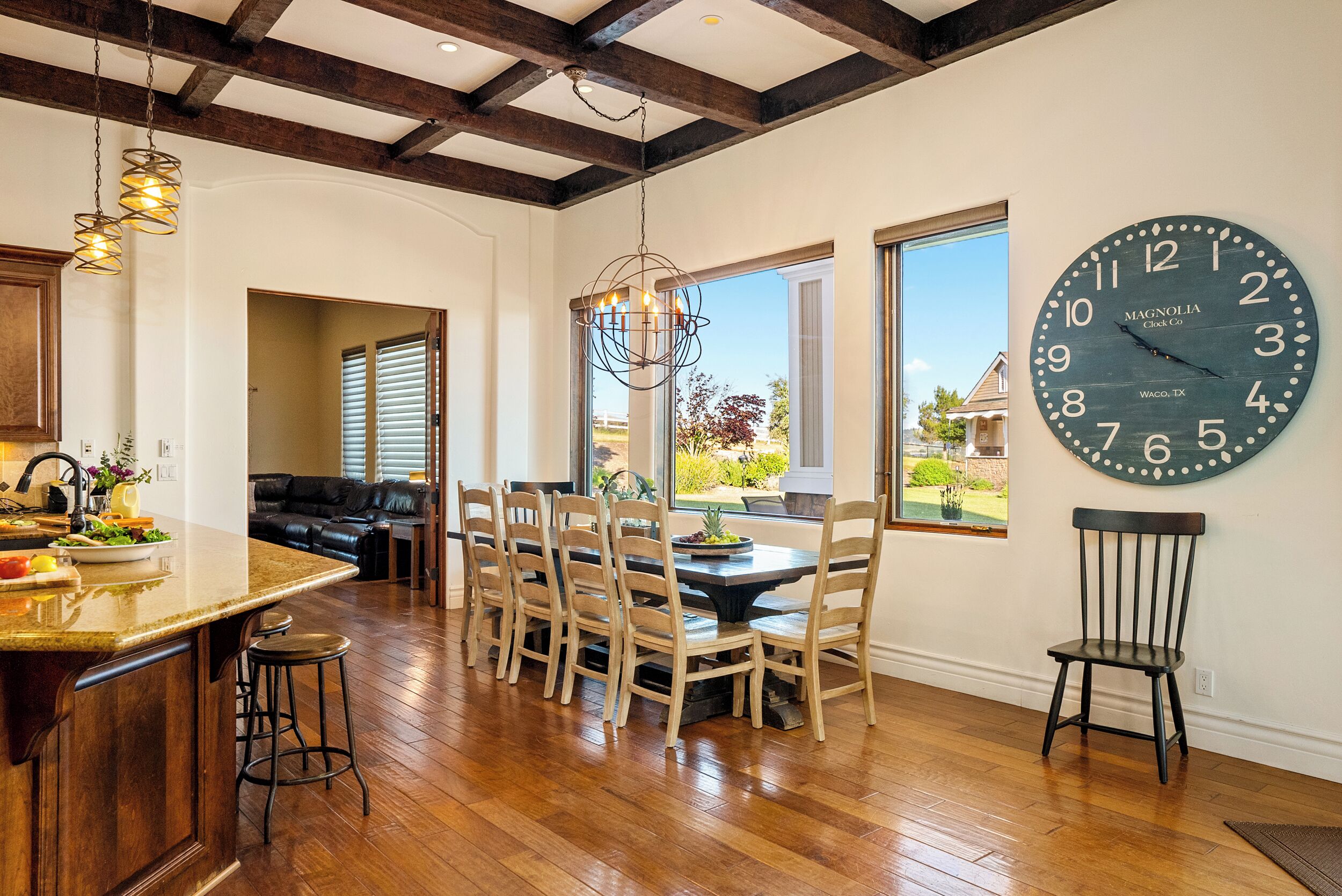 Une cuisine spacieuse avec un plancher en bois, un coin repas avec six chaises, une grande horloge murale et une vue sur l’extérieur grâce à de grandes fenêtres.