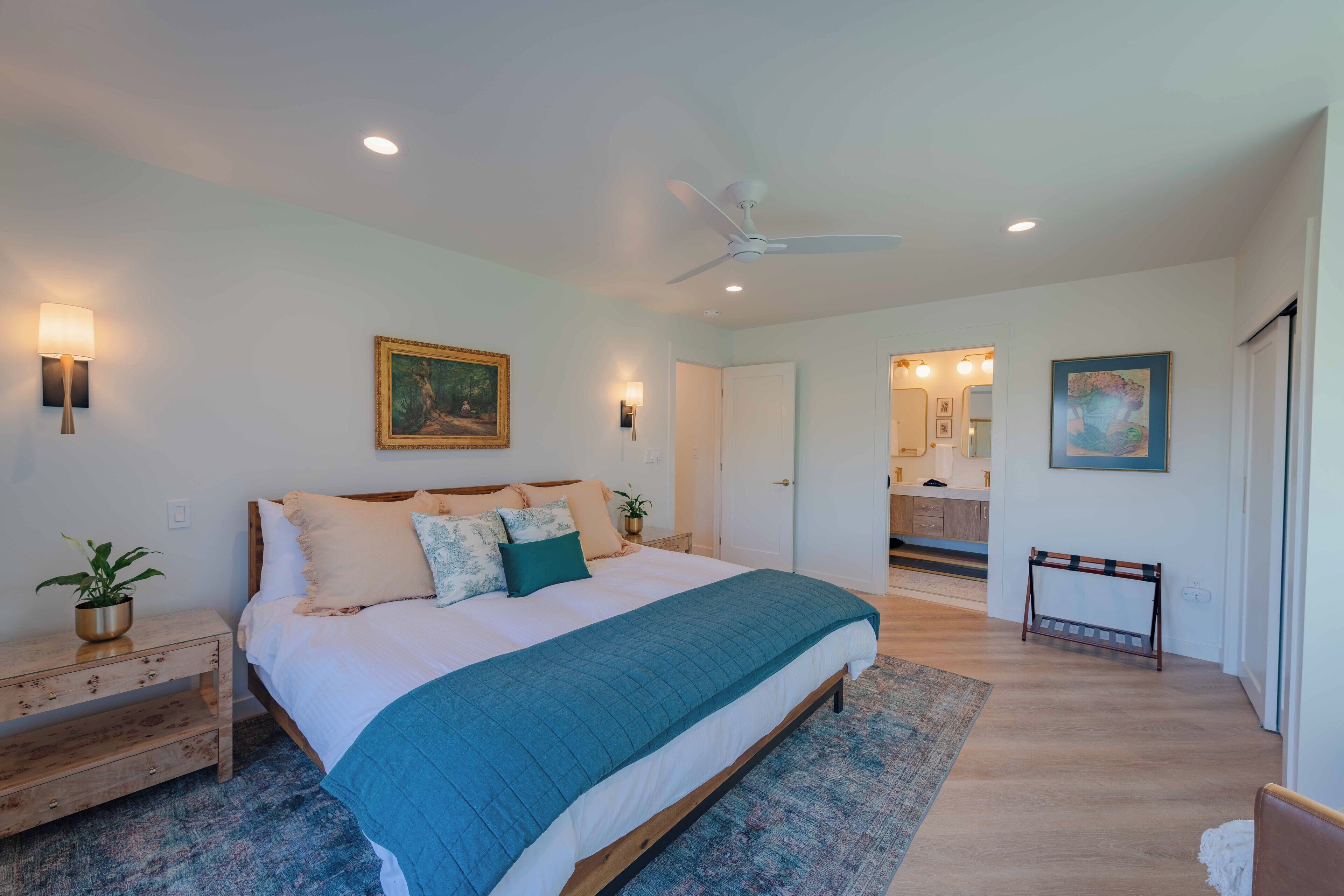 A bedroom with a large bed, bedside tables, a ceiling fan, and a view into the kitchen.