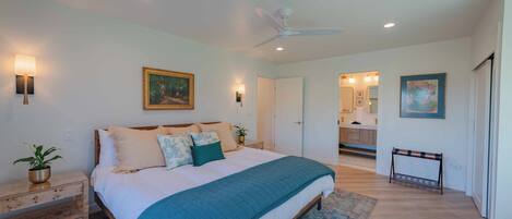 A bedroom with a large bed, bedside tables, a ceiling fan, and a view into the kitchen.