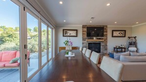 A spacious living room with a long wooden dining table, a fireplace, and large windows offering a view of the outdoors. - Bella Vita (Paso Robles)