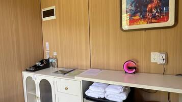 Habitación de hotel compacta con pared de paneles de madera, un escritorio blanco con una computadora, un refrigerador pequeño, un banco y un cuadro de un vaquero y un caballo.