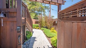 A wooden gate with a keypad lock, a paved pathway, and a well-maintained garden with various shrubs and a small tree. - Moonglow Cottage (Cambria)