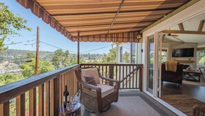 A balcony with a wicker chair, a small table with a bottle and glass, and a view of a town. - Moonglow Cottage (Cambria)