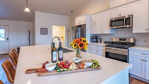 A modern kitchen with white cabinets, a large island with a fruit and cheese board, a sink, and stainless steel appliances. - Blue Moon Escape (Paso Robles)