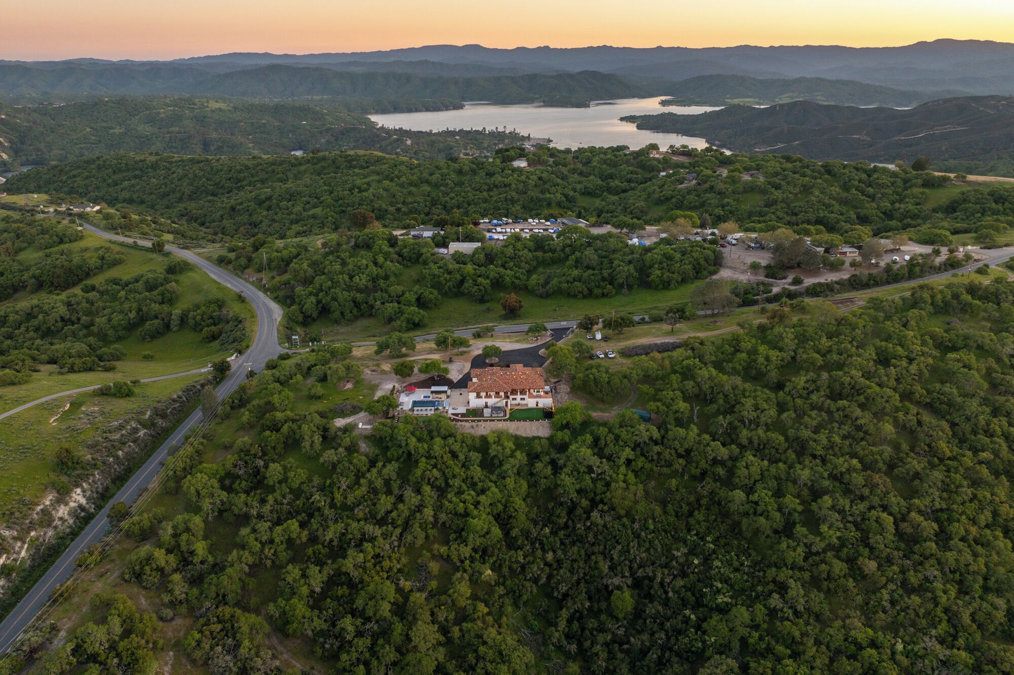 An aerial view of a rural area with a large house surrounded by dense forest, a winding road, and a lake in the background.