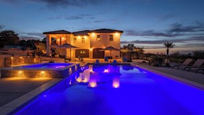 A swimming pool with blue lighting, a house with a tiled roof, and outdoor seating.