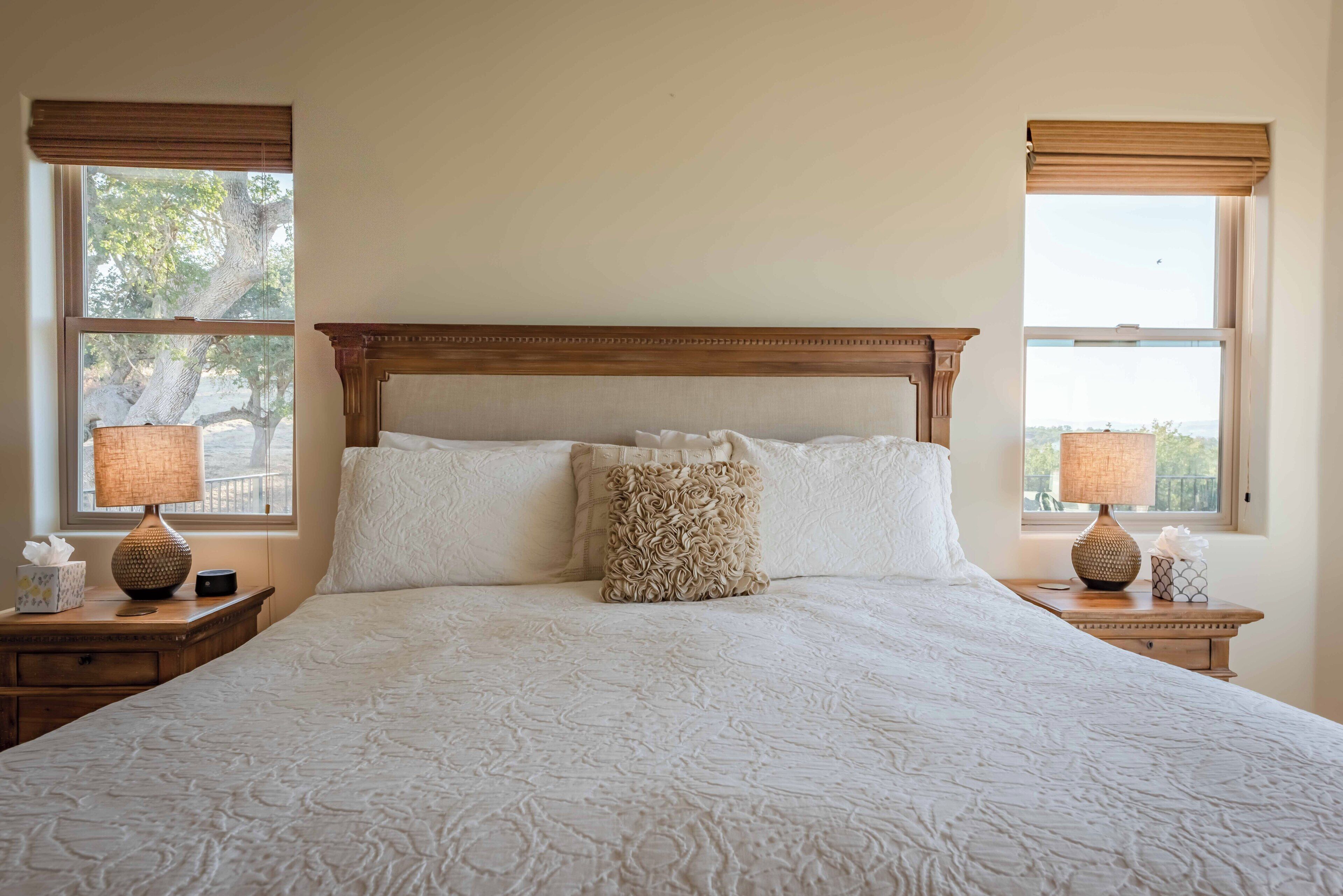 A large bed with a wooden headboard, two bedside lamps, and a view of trees through the windows.