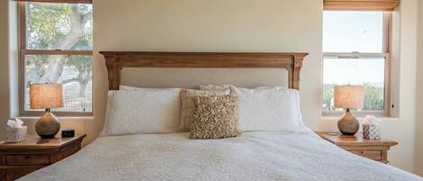 A large bed with a wooden headboard, two bedside lamps, and a view of trees through the windows.