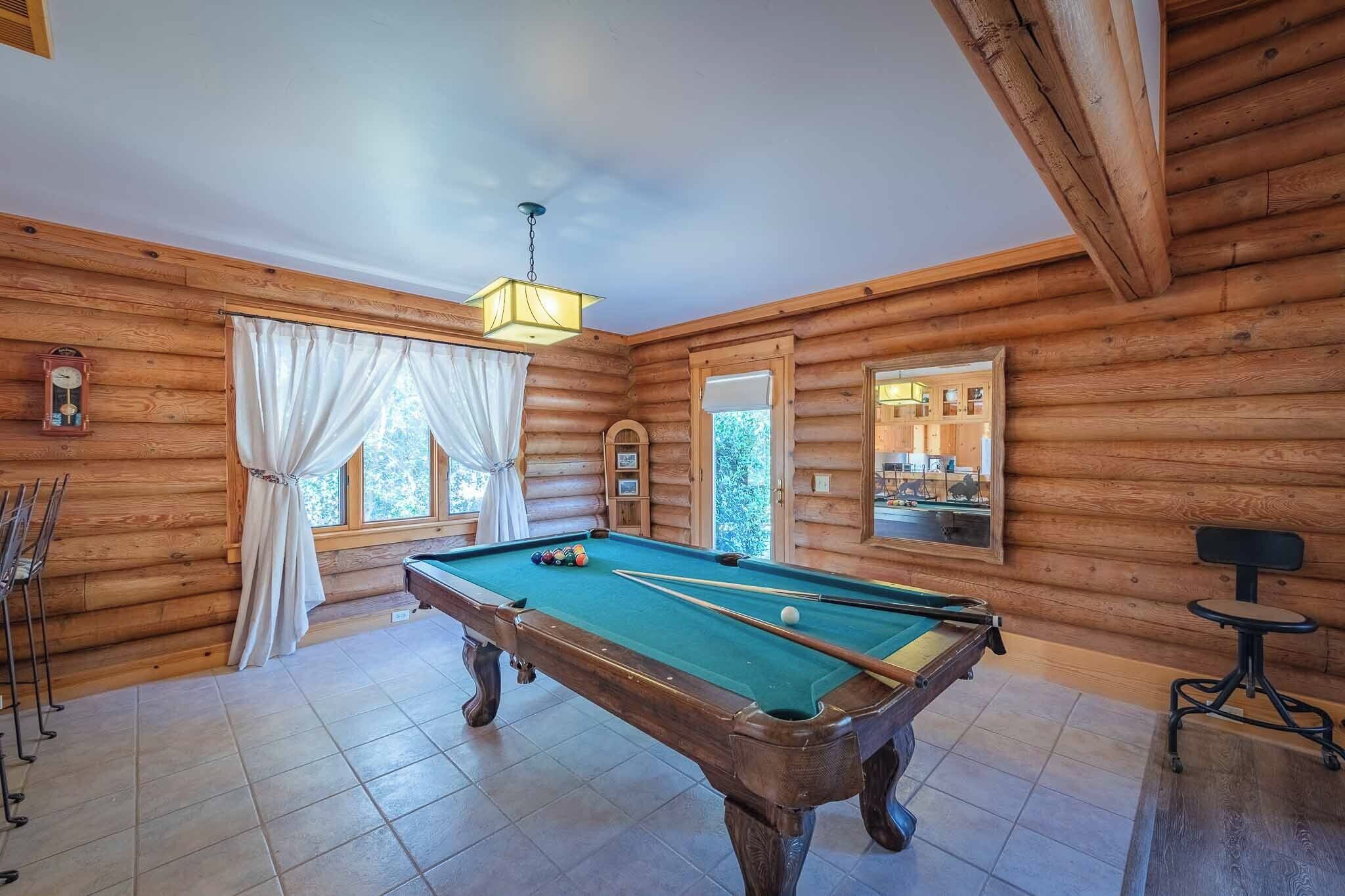 Une table de billard dans un chalet en rondins, agrémentée d’un lustre, d’une horloge et d’une fenêtre avec des rideaux.