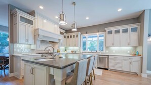 A spacious kitchen with white cabinetry, a large island with bar stools, and a view of the outdoors through large windows. - Sycamore Ranch (Templeton)