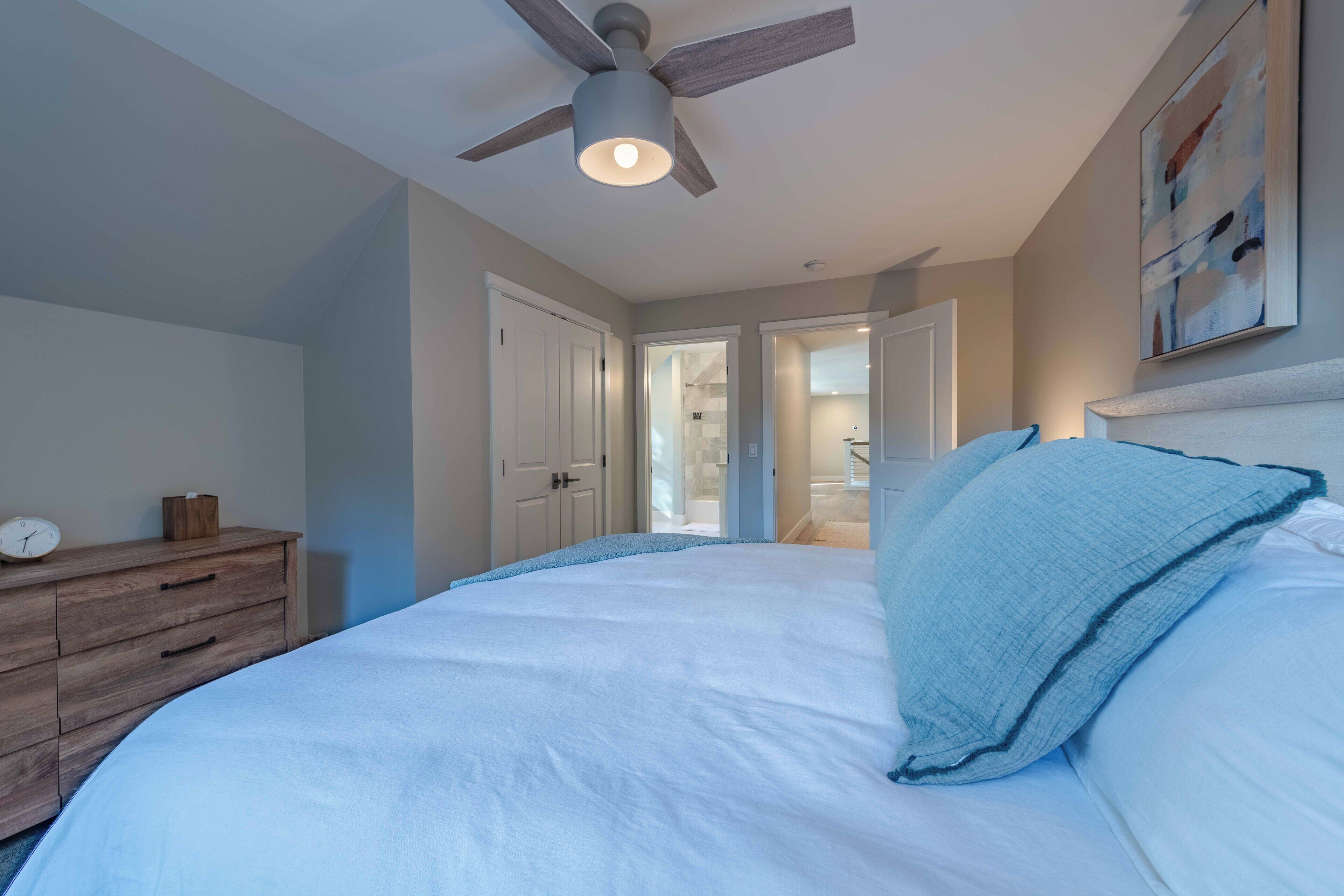 A bedroom with a bed, a wooden dresser, a ceiling fan, and a door leading to another room.