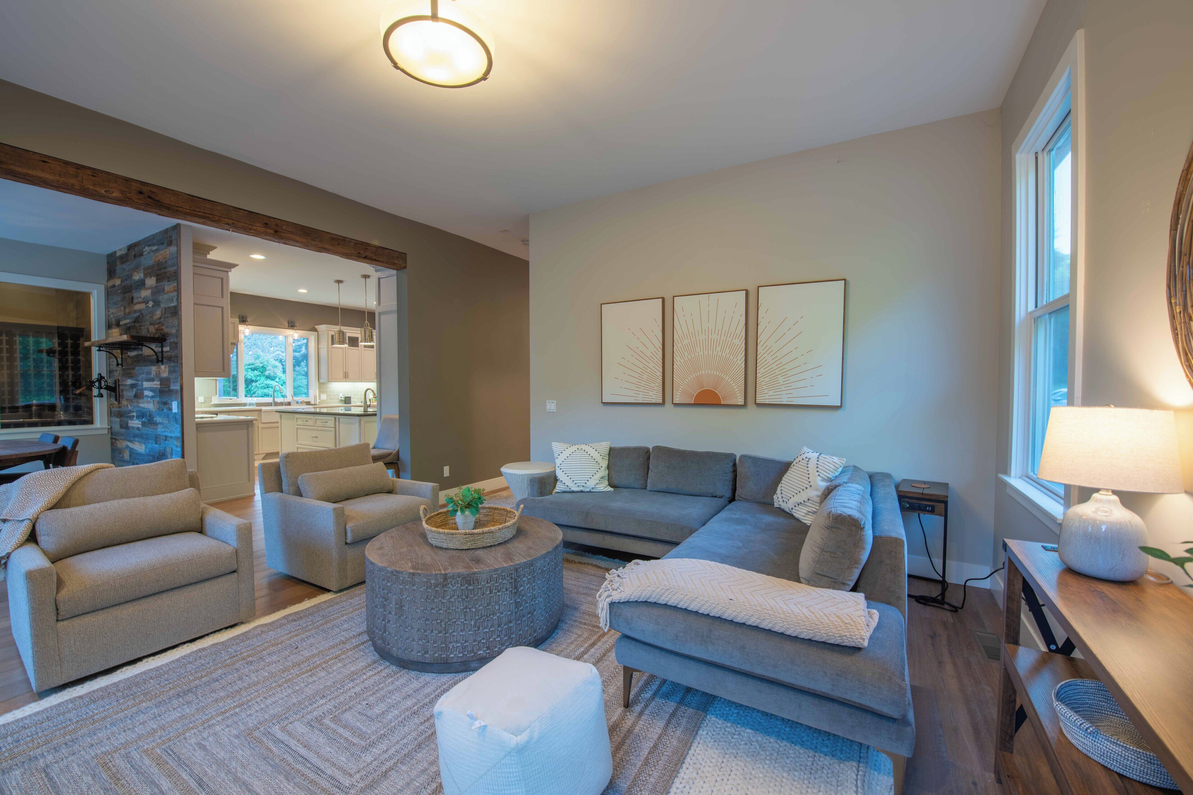 A modern living room with a grey sofa, two armchairs, a coffee table, and a fireplace. There is a kitchen in the background with white cabinets and a stone accent wall.