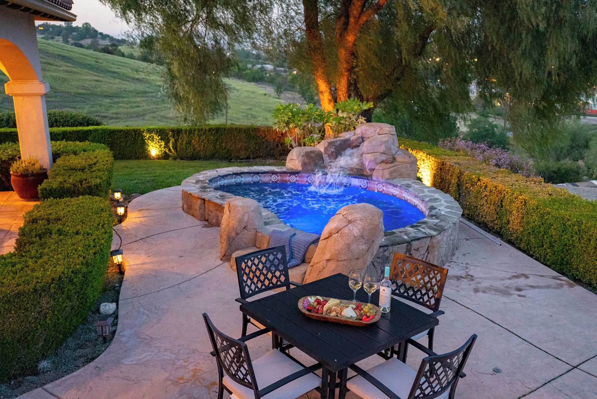 A hot tub surrounded by a landscaped garden with a table set for a meal.