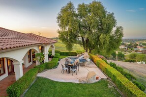 A patio with a hot tub, a dining table, and chairs, surrounded by well-manicured hedges and a large tree.