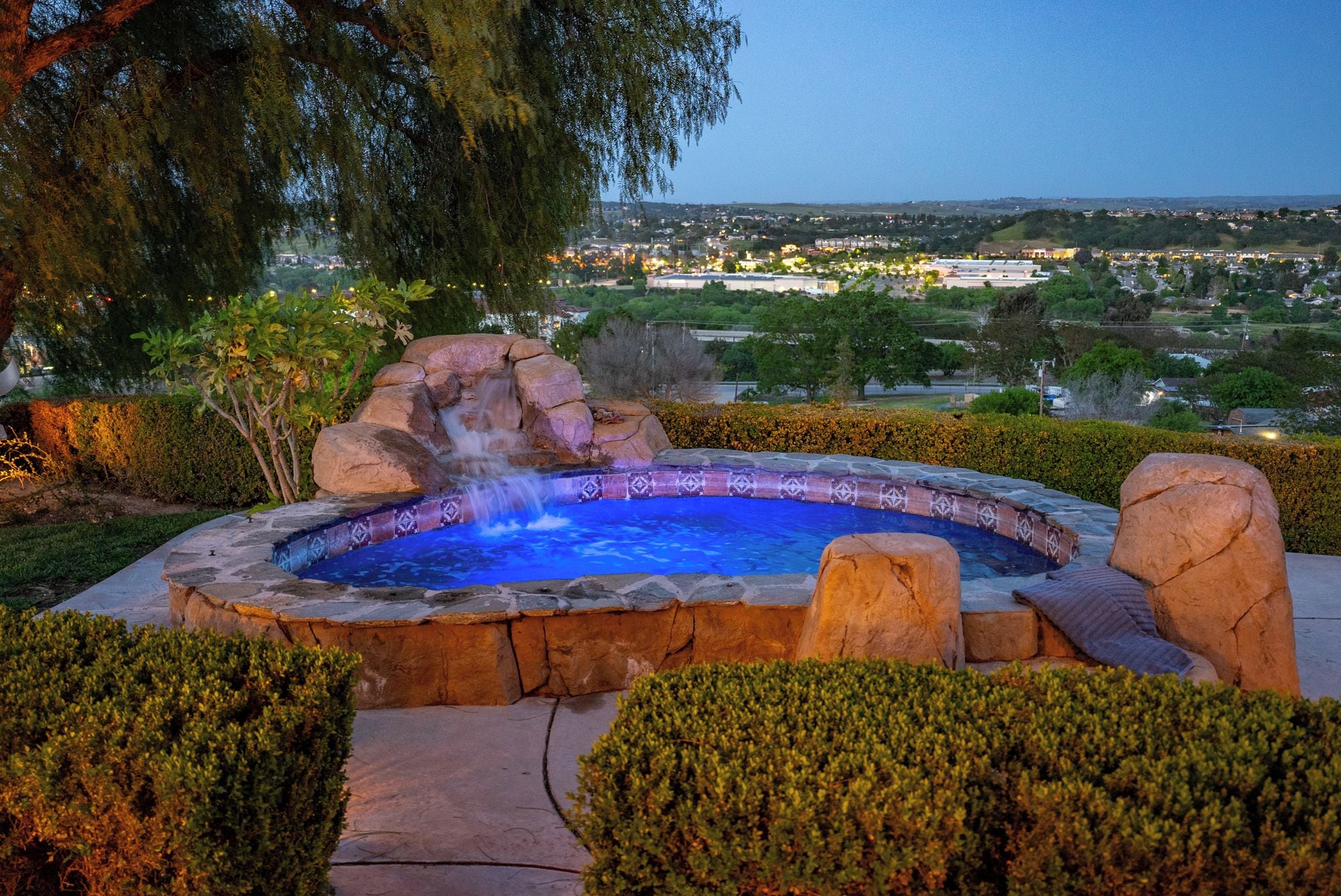 Un jacuzzi con cascada, rodeado de setos cuidados y vista a una zona residencial.