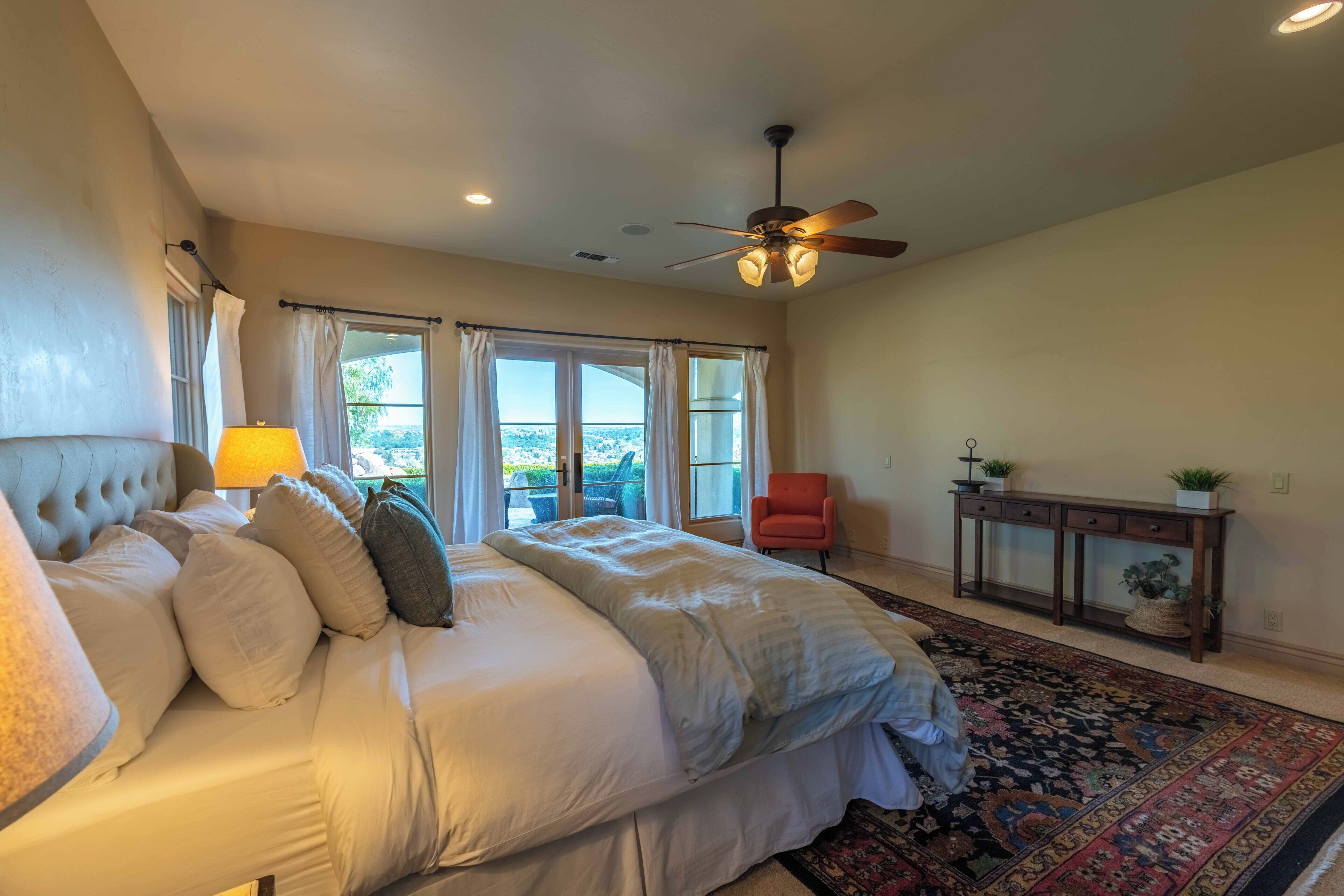 A bedroom with a bed, a ceiling fan, a window with curtains, a chair, a console table with plants, and a patterned rug.
