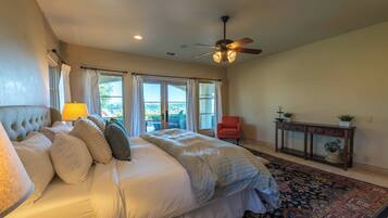 A bedroom with a bed, a ceiling fan, a window with curtains, a chair, a console table with plants, and a patterned rug.