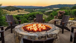 A stone fire pit with chairs around it, set against a backdrop of a sunset over a hilly landscape. - Paso Paradiso (Paso Robles)