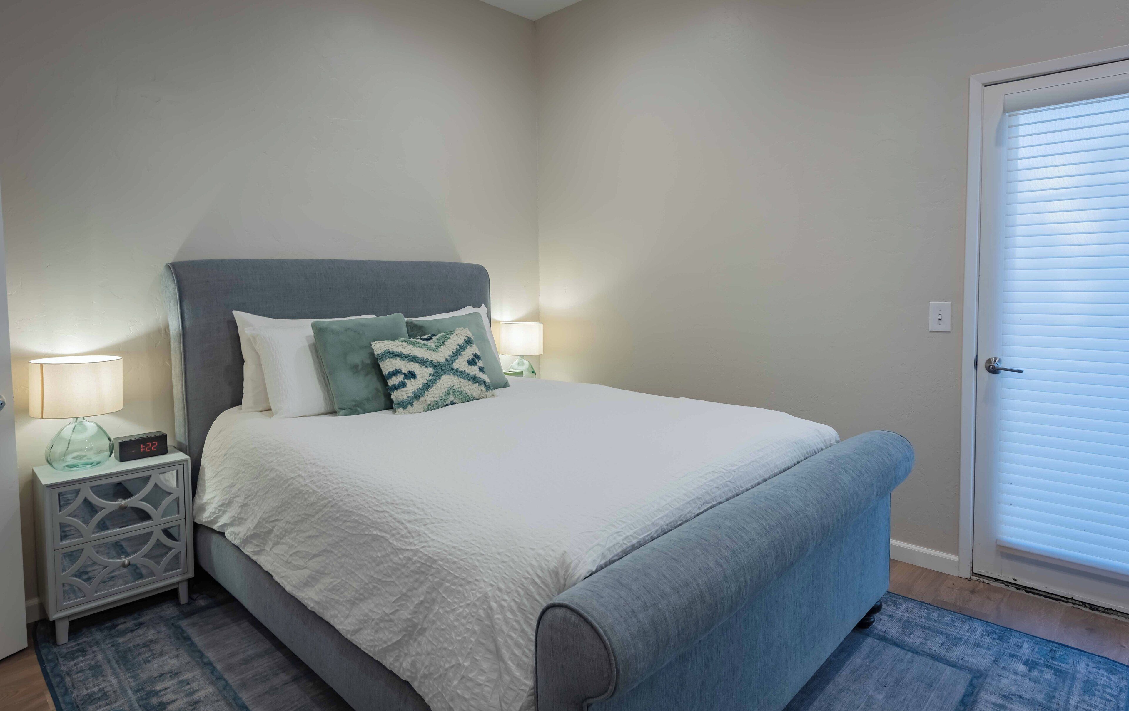 Een net opgemaakt bed met een grijze hoofdbord, witte beddengoed en een blauw sierkussen. Er staat een nachtkastje met een lamp en een digitale klok. De kamer heeft een eenvoudig, schoon ontwerp met een deur en een raam met zonwering.