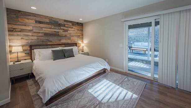 Une chambre à coucher avec une tête de lit en bois, un grand lit, des lampes de chevet et une vue sur un patio extérieur, accessible par une porte-fenêtre coulissante.