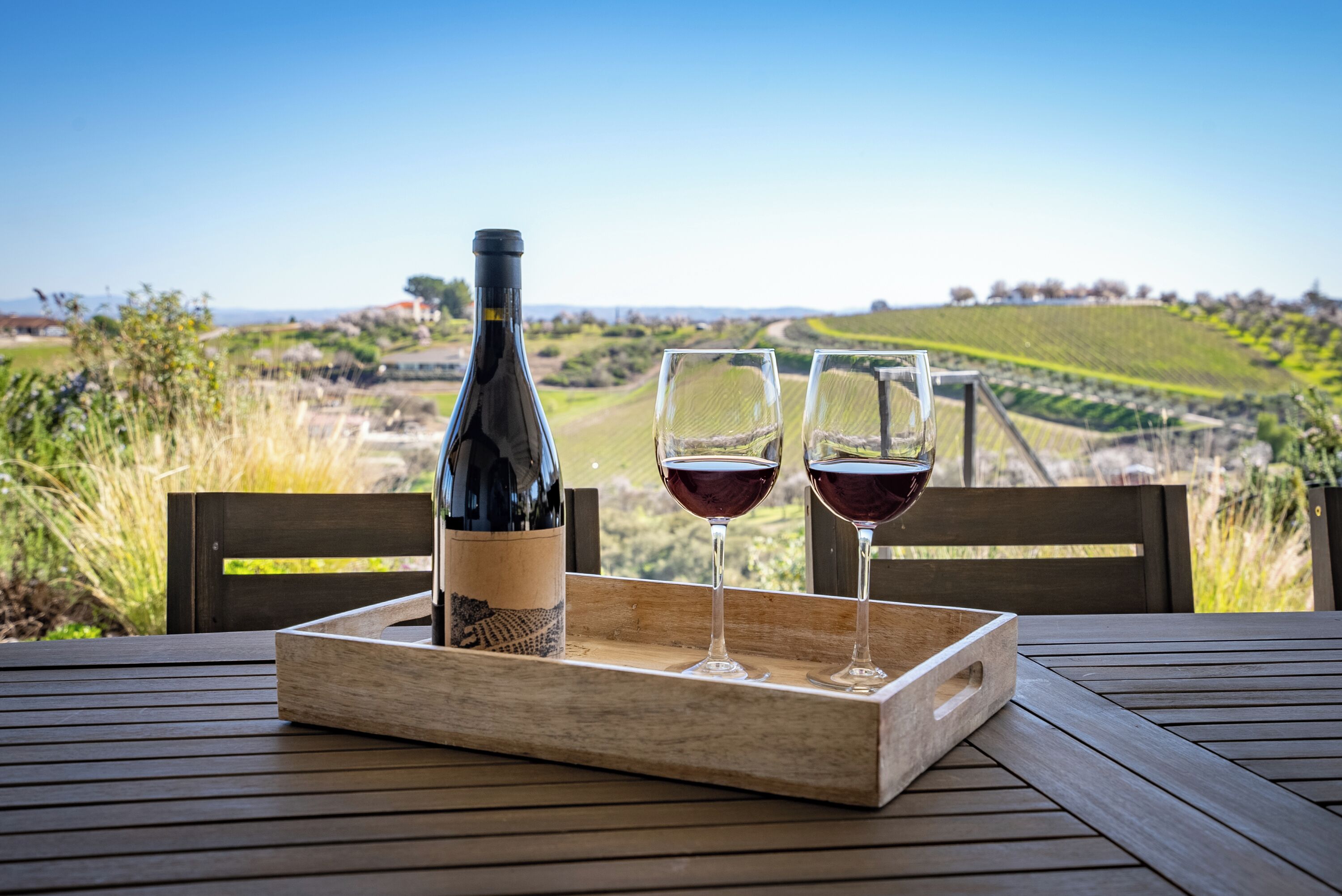 Une table en bois, sur laquelle se trouvent une bouteille de vin et deux verres, le tout posé sur un plateau, avec une vue sur un vignoble.