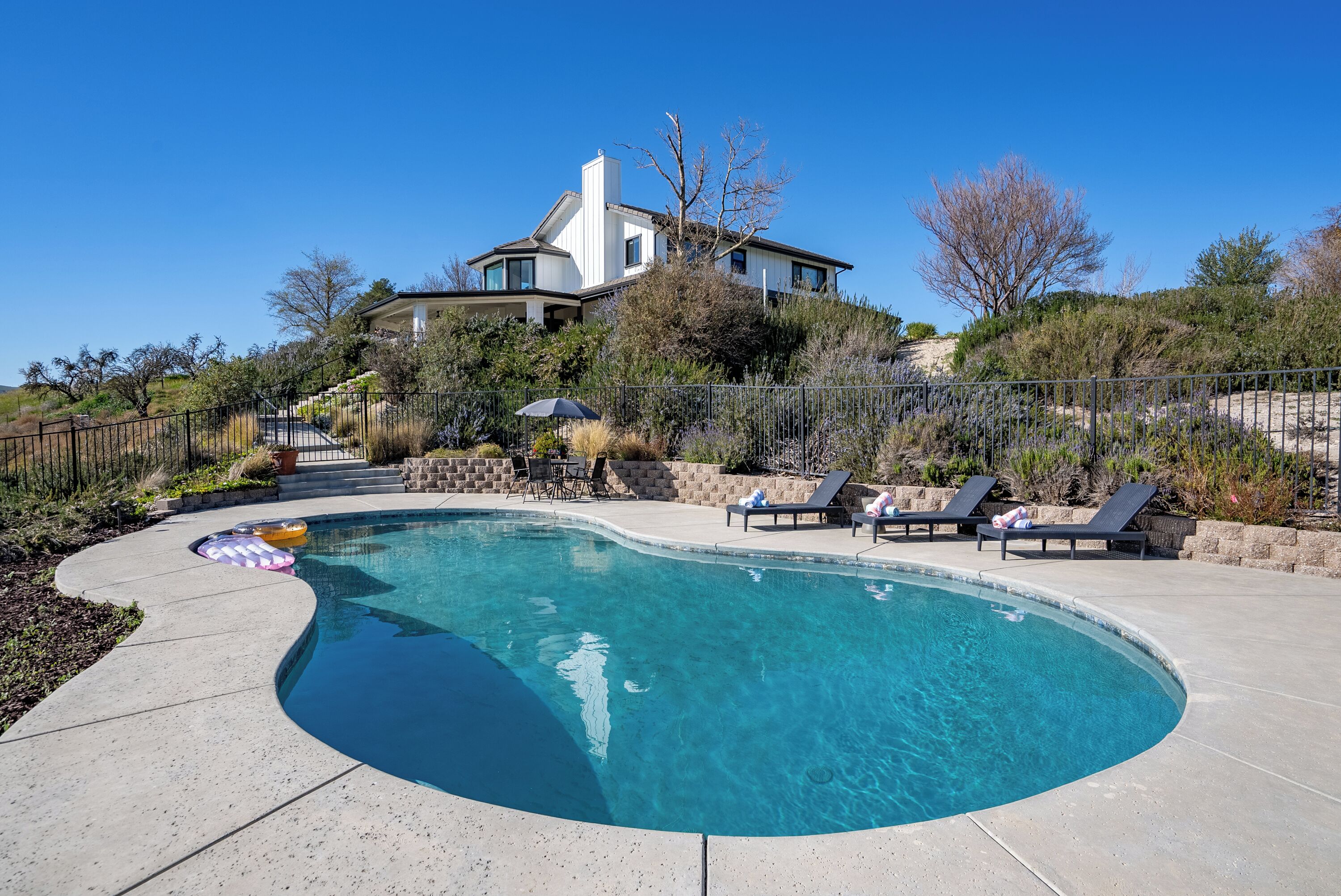 Une piscine avec des chaises longues et un parasol, entourée d’un jardin aménagé et d’une maison en arrière-plan.