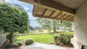 A covered porch with a concrete floor, surrounded by greenery and a view of a well-maintained lawn and trees. - Golfer's Getaway (Nipomo)