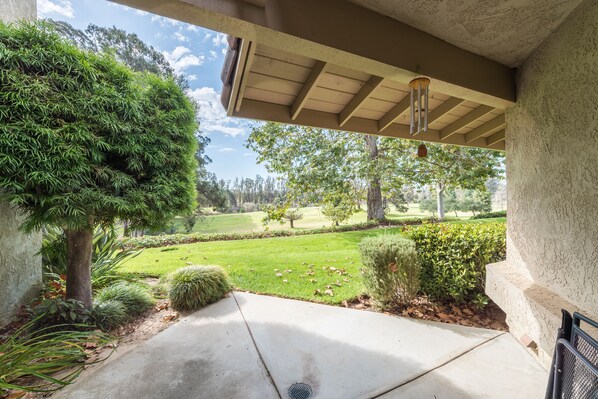 A covered porch with a concrete floor, surrounded by greenery and a view of a well-maintained lawn and trees. - Golfer's Getaway (Nipomo)