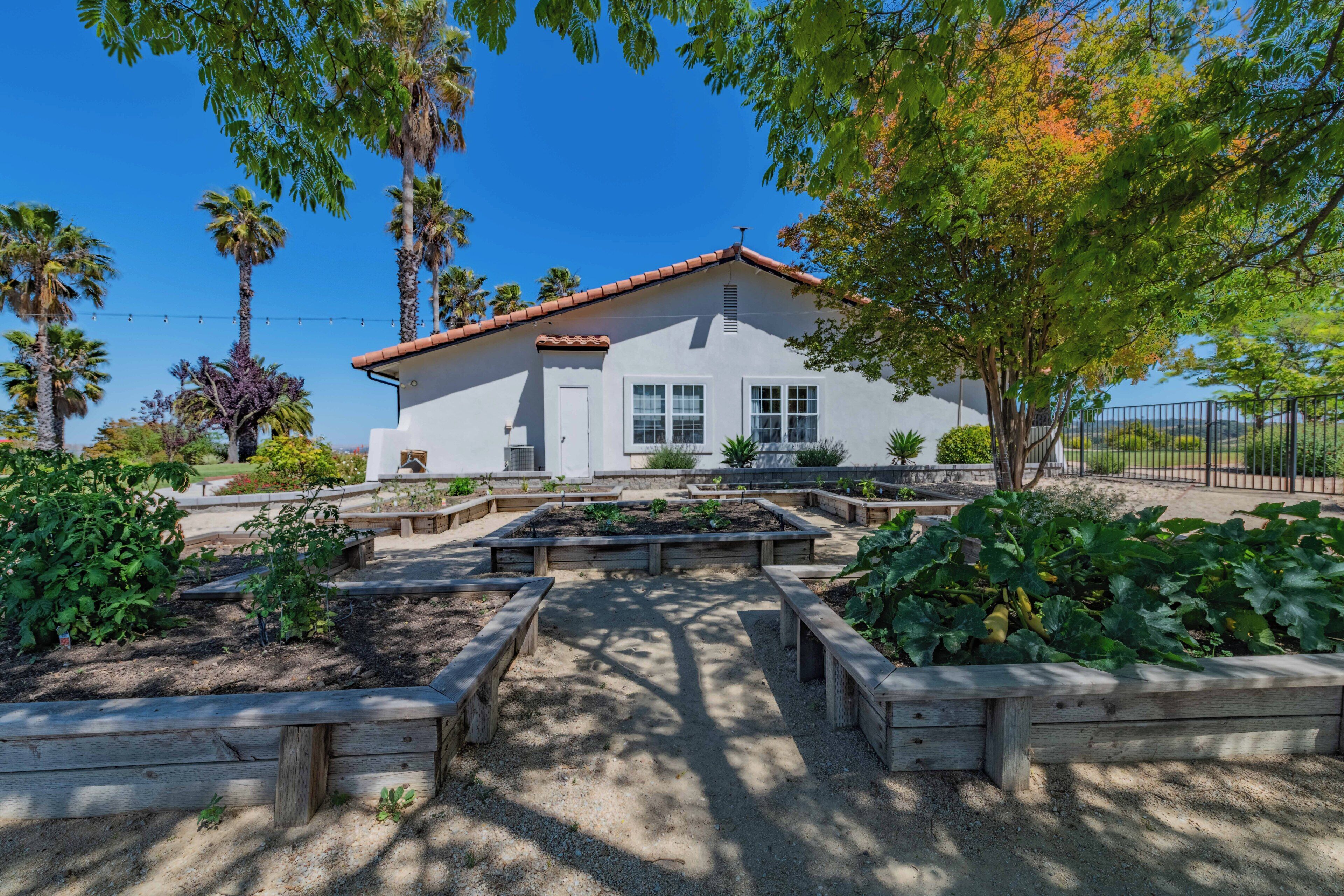 A white building with a red roof, surrounded by raised garden beds and palm trees.