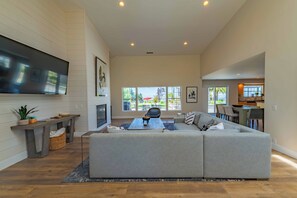 Un salón amplio con un televisor de pantalla plana grande, un sofá modular gris, una mesa de centro de madera y una zona de comedor con mesa y sillas.