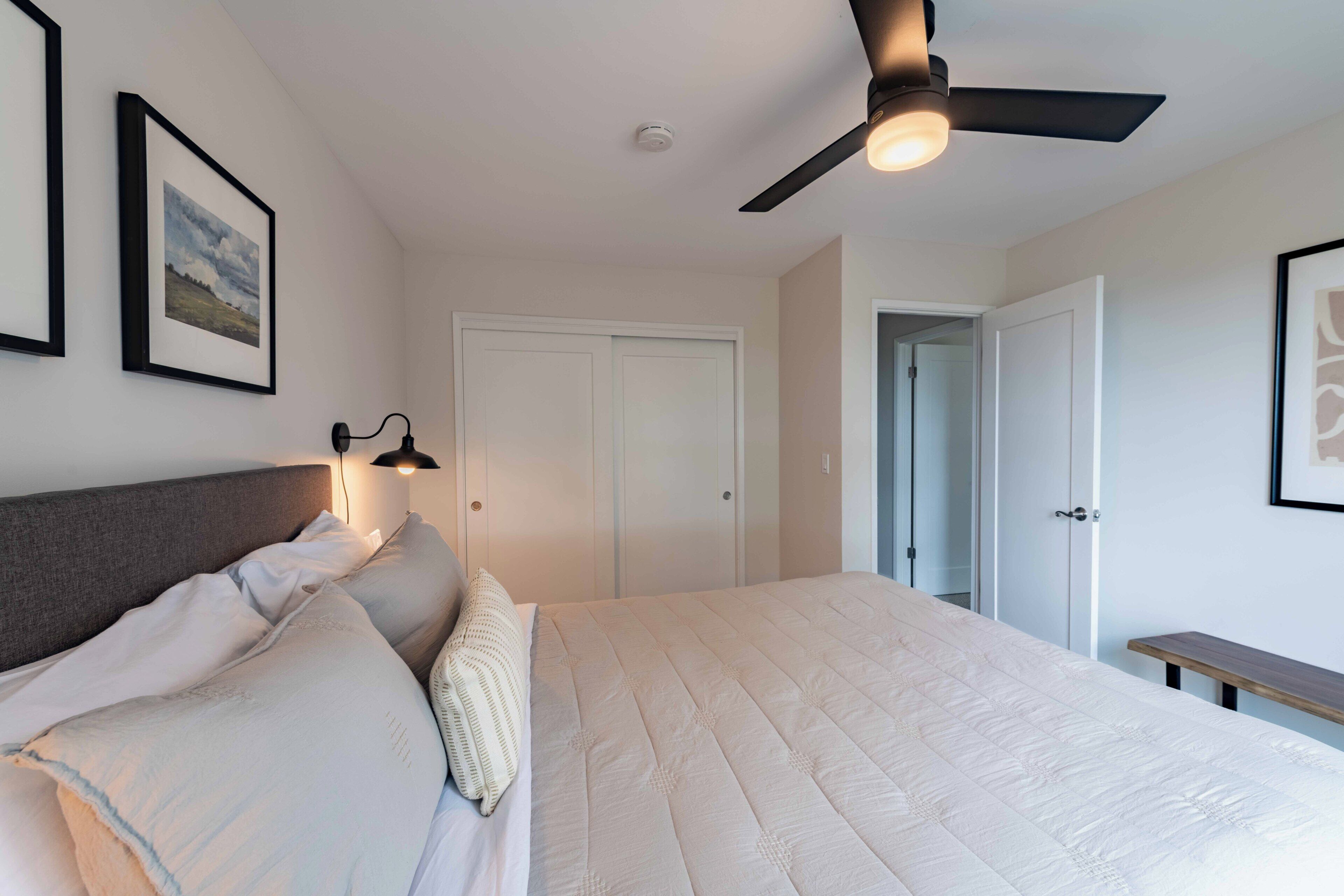 A bedroom with a bed, a ceiling fan, a bedside lamp, and framed pictures on the wall.