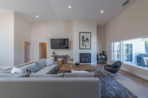 A modern living room with a sectional sofa, a fireplace, a flat-screen TV, and large windows.