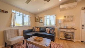 A living room with a sectional sofa, a chair, a coffee table, and a desk with a lamp. There are windows, a ceiling fan, and an air conditioning unit. - Hidden Acre - Bird House (Paso Robles)