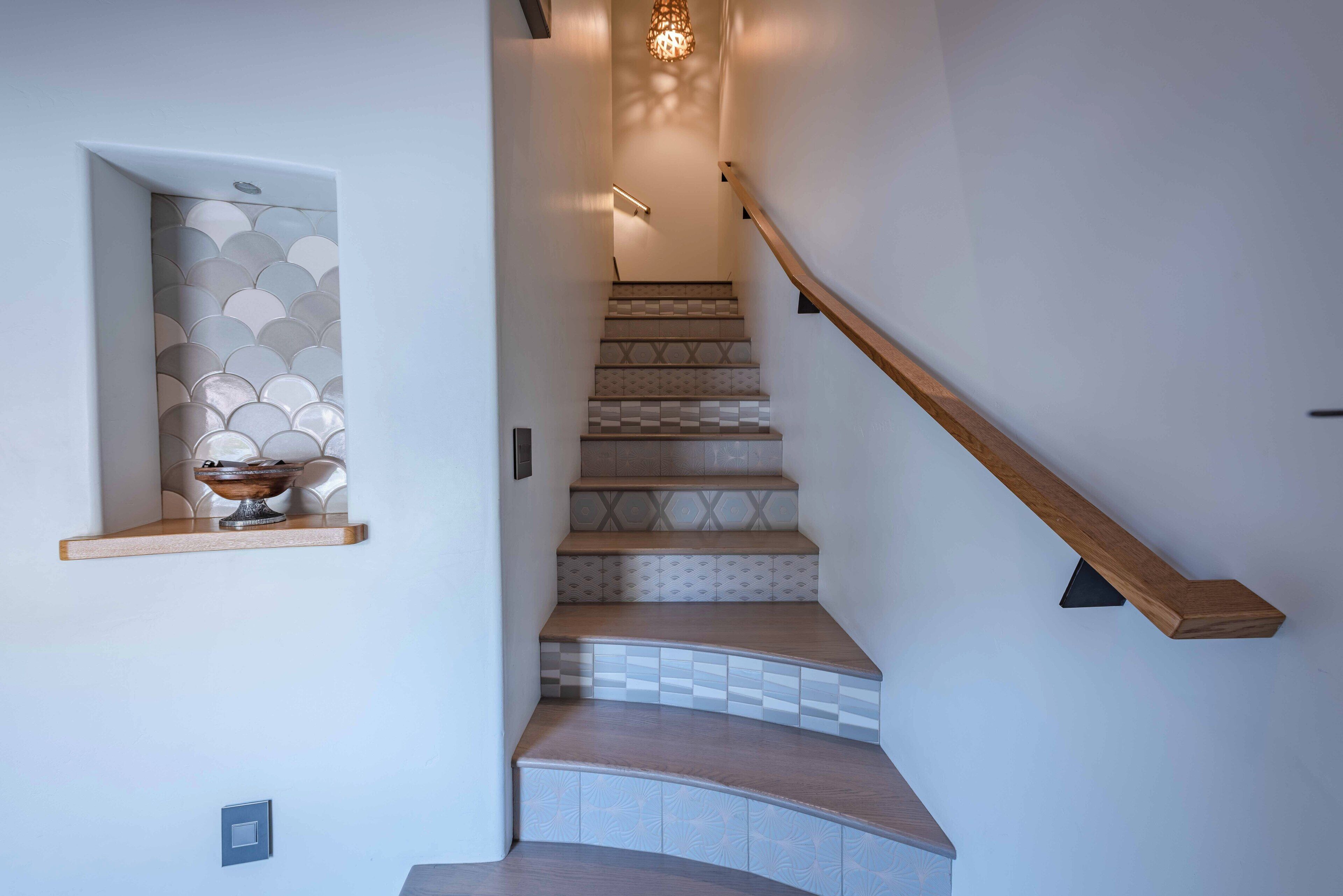 Una escalera con baldosas estampadas, barandilla de madera y un estante de pared con un objeto decorativo.