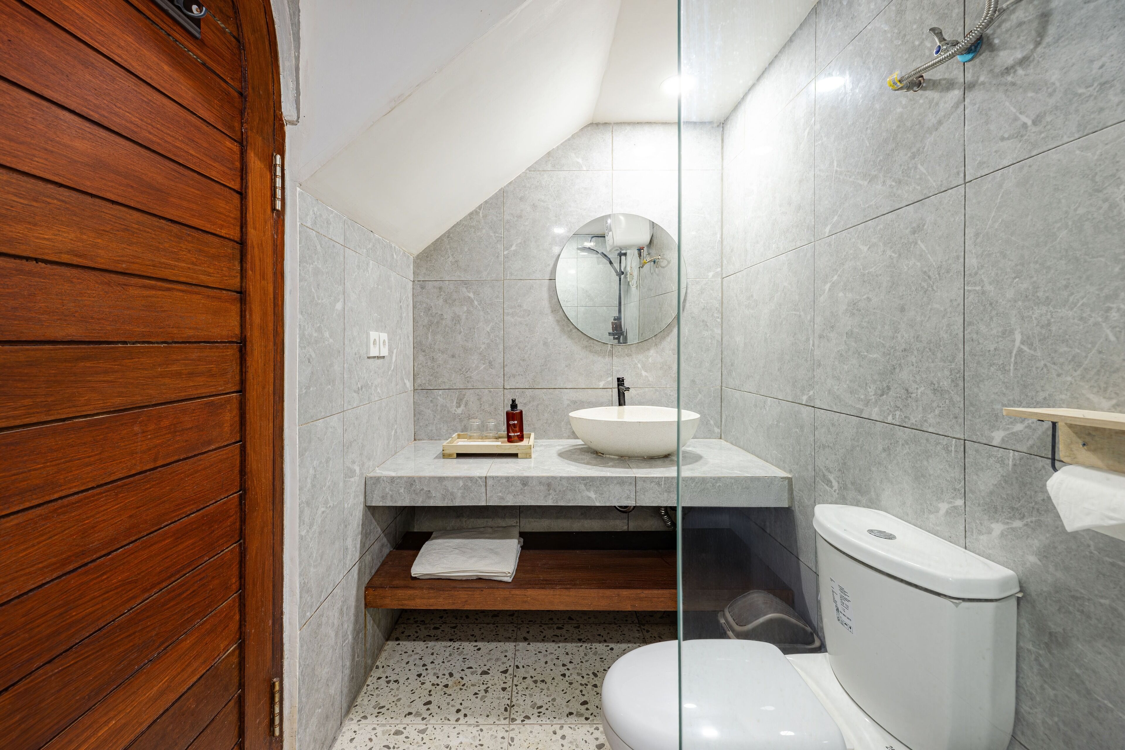 Um banheiro com porta de madeira, espelho redondo, pia branca, vaso sanitário e prateleira de madeira.