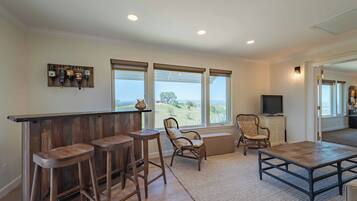A spacious living area with a wooden bar, two cushioned chairs, a television, and large windows offering a view of the outdoors.