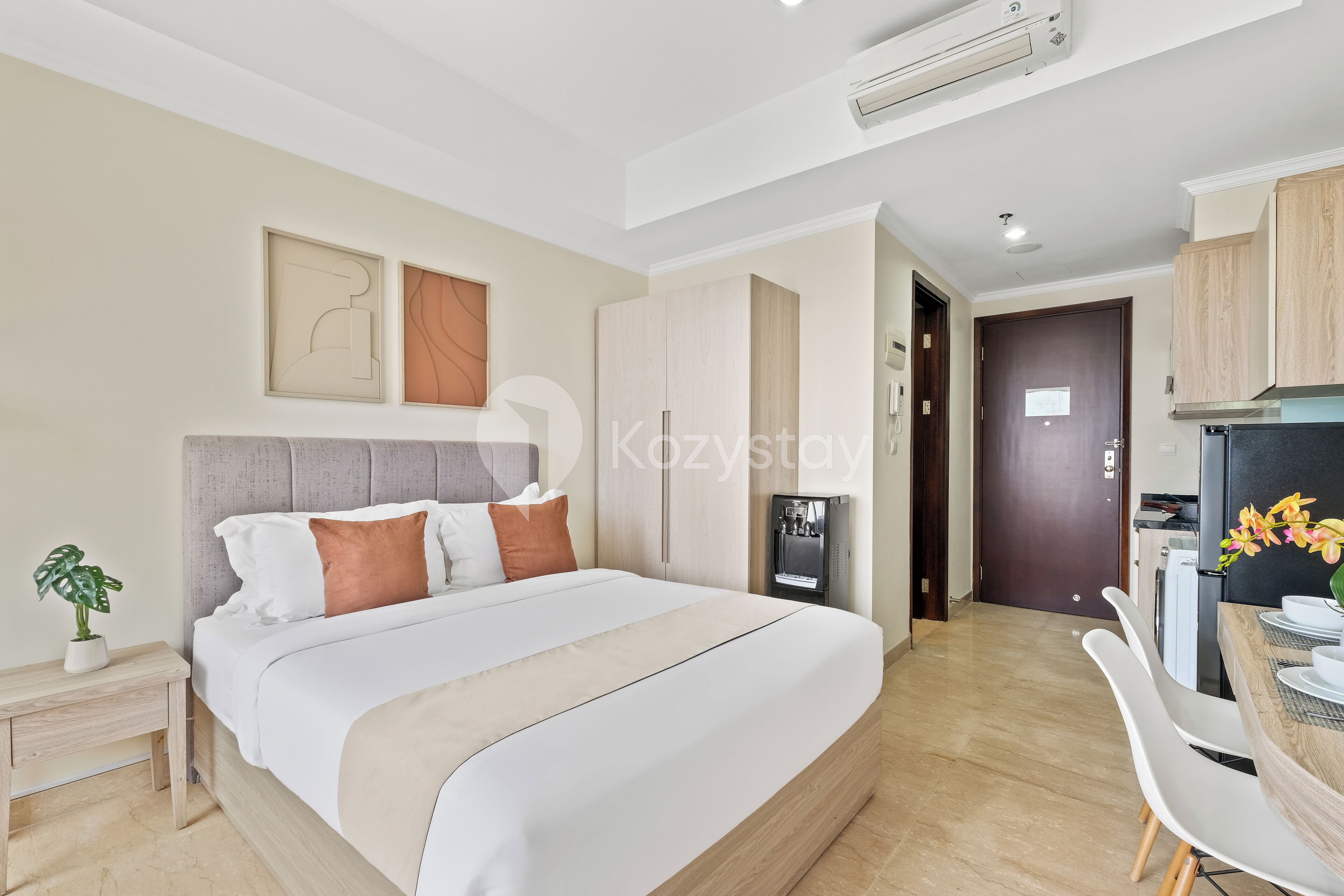 A modern hotel room with a large bed, a small bedside table with a plant, a desk with a chair, and a kitchenette area.