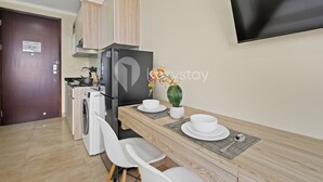 A compact kitchen with a washing machine, a refrigerator, wooden cabinets, and a dining area with a table set for two. - Brenna by Kozystay - Menteng (Jakarta)