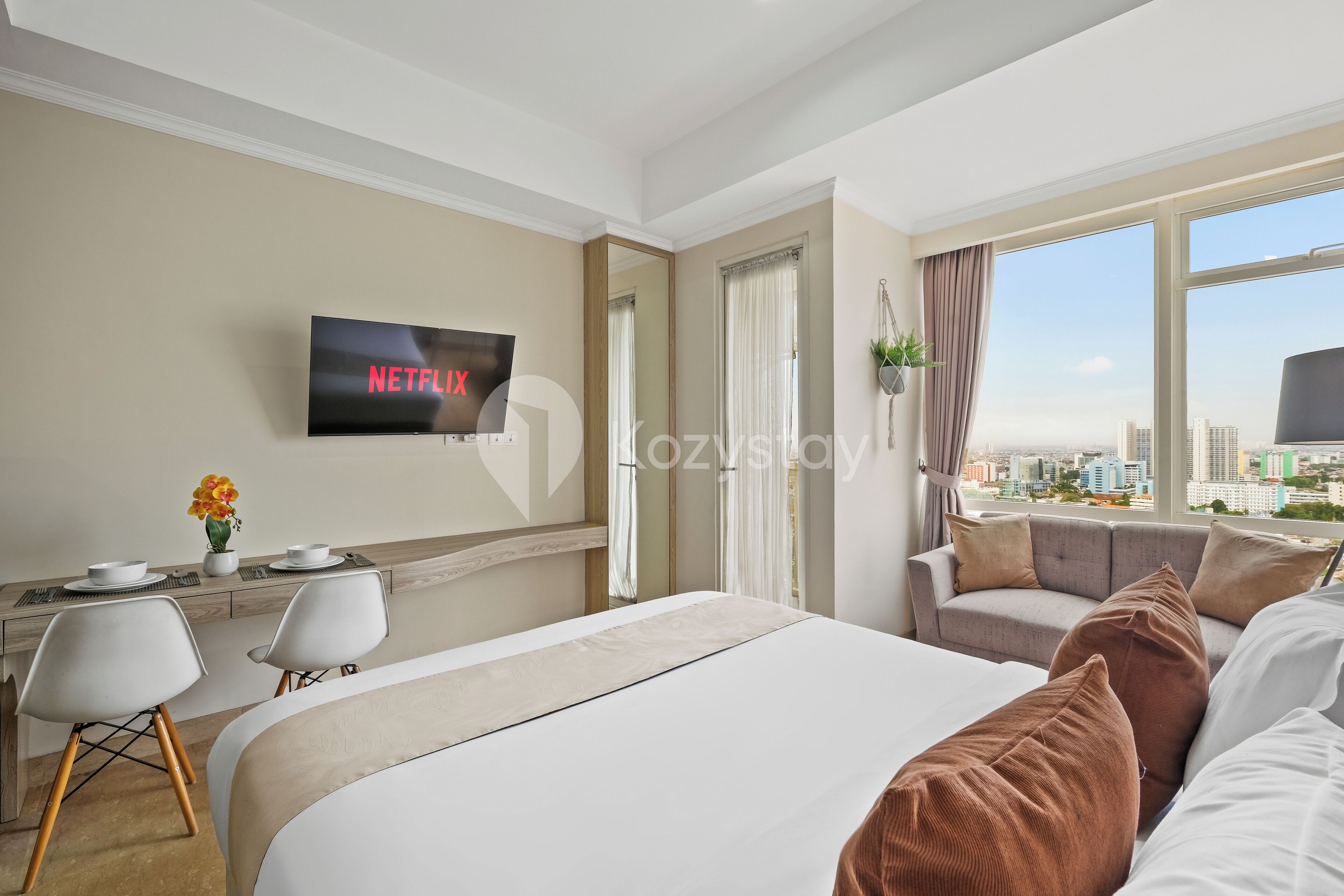 Habitación de hotel moderna con una cama grande, un escritorio con una televisión mostrando 'NETFLIX', un sofá y vistas a la ciudad a través de amplios ventanales.
