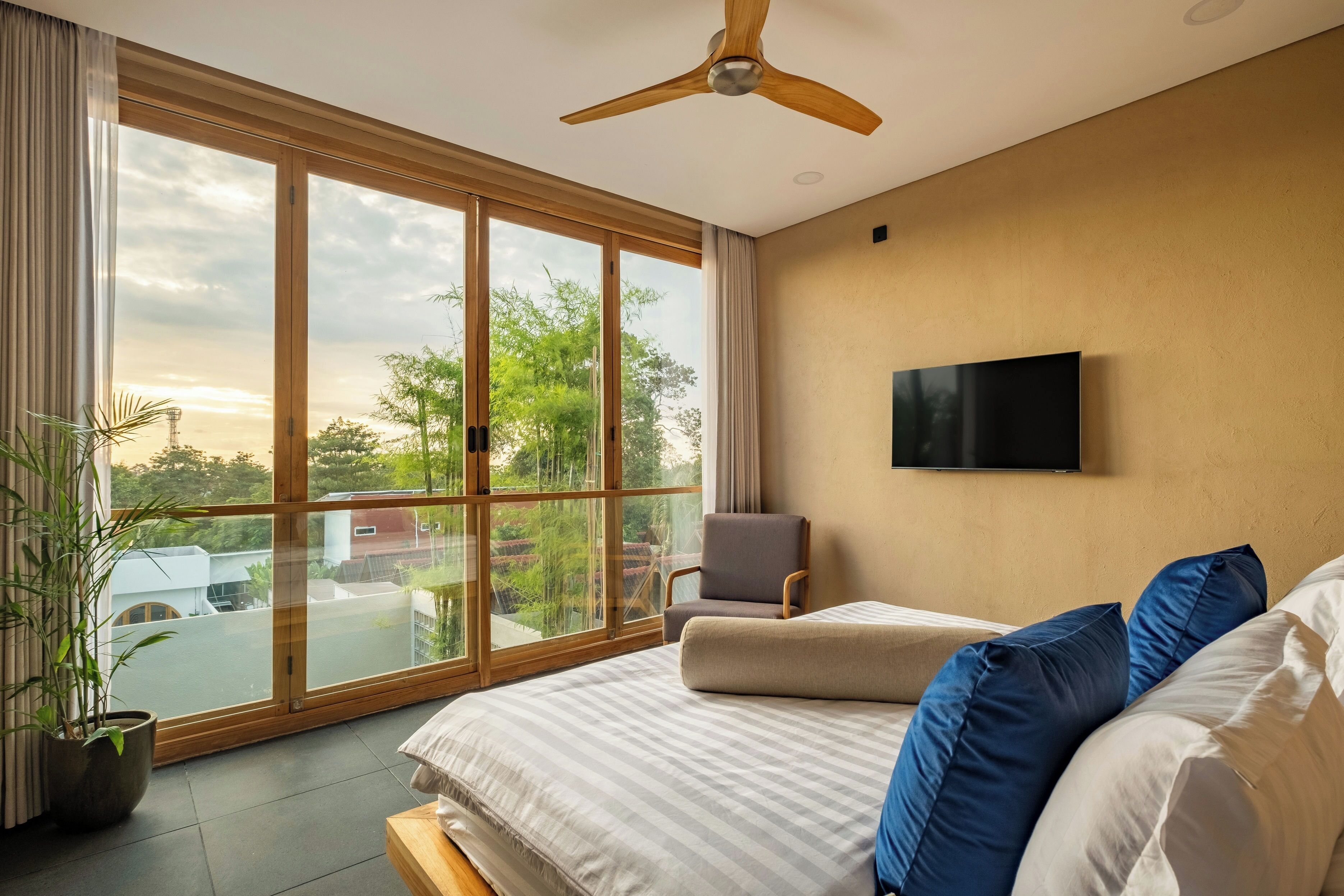 A bedroom with a large sliding glass door, a ceiling fan, a flat-screen TV, a bed with blue and white pillows, and a potted plant.