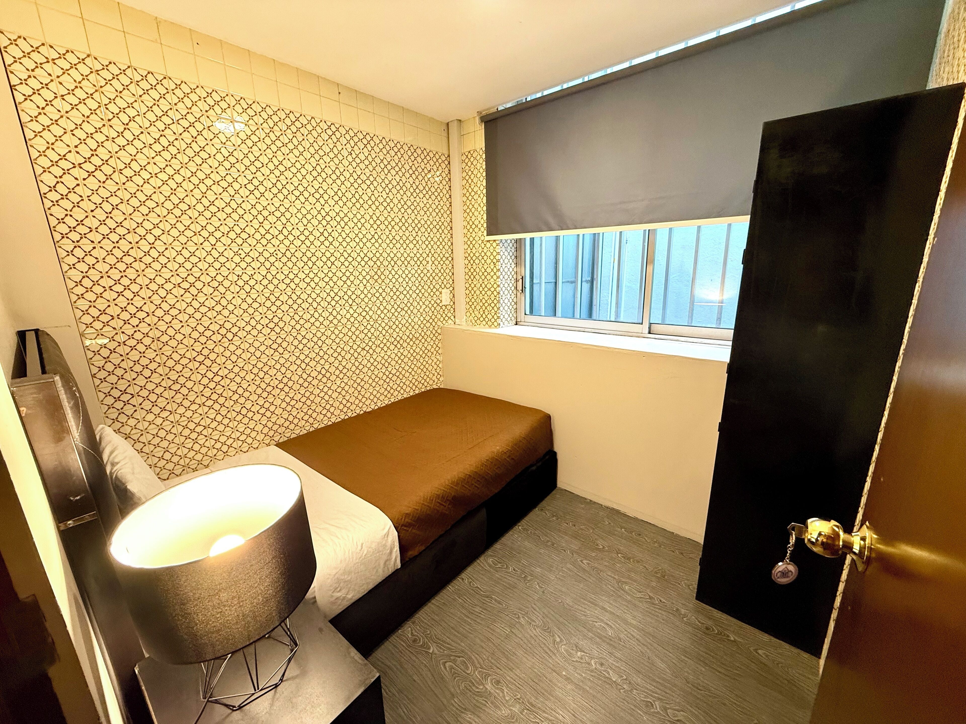 A small, compact room with a bed, a bedside table with a lamp, a window with blinds, and a patterned wall.