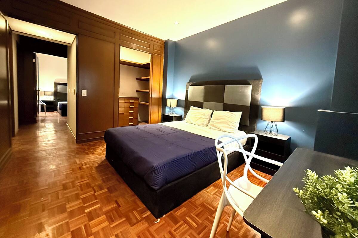 A hotel room with a bed, bedside table, lamp, and a view of the hallway.