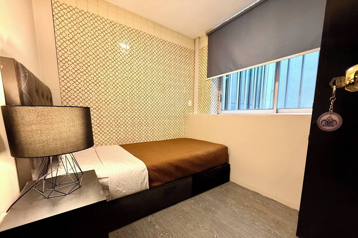 A small, modern bedroom with a bed, a desk with a lamp, a patterned wall, and a window with blinds.