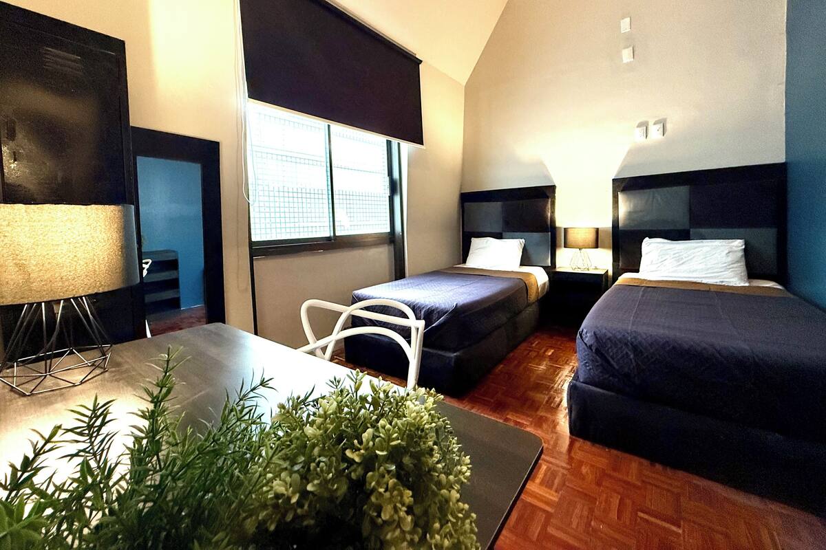 A hotel room with two beds, a desk, a chair, a TV, and a window with blinds.
