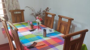Une table à manger recouverte d’une nappe colorée, entourée de chaises en bois. Un vase rempli de fleurs et une bouteille sont posés sur la table.