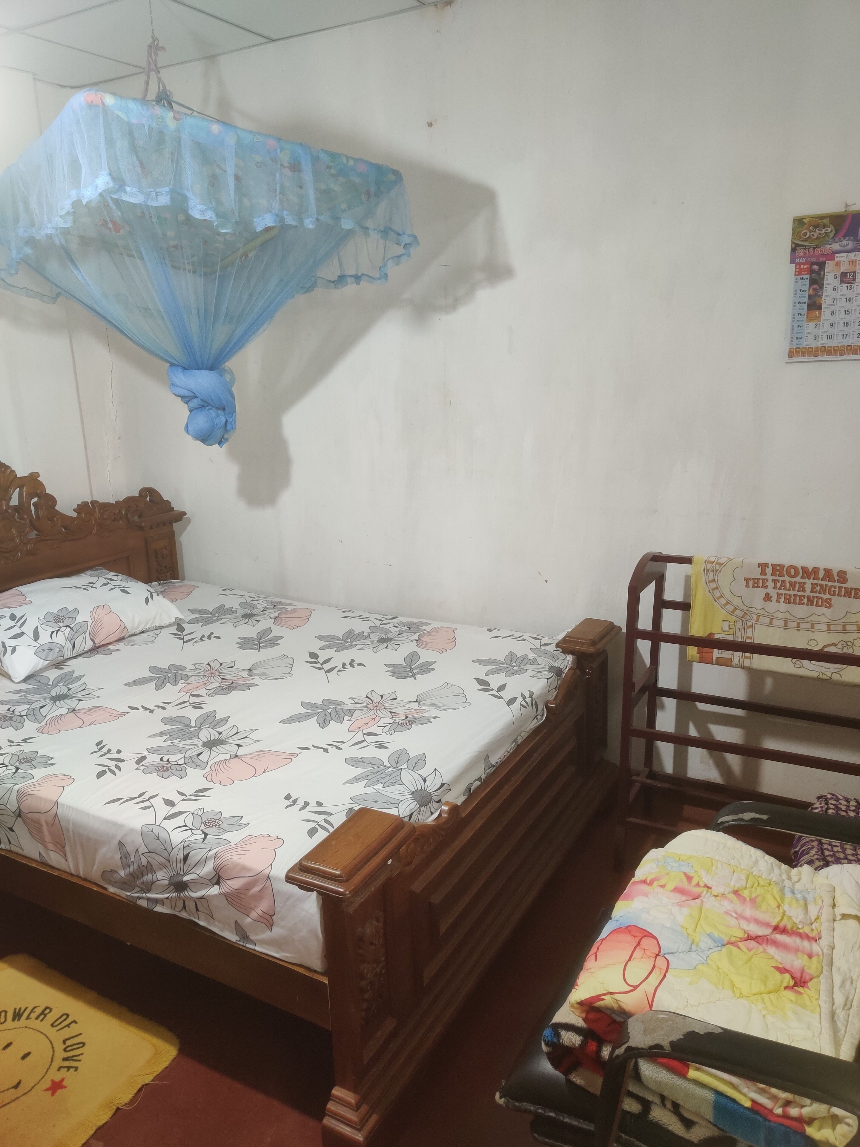 A bedroom with a bed, a wooden headboard, a small table, a chair, a mosquito net, and a calendar on the wall.
