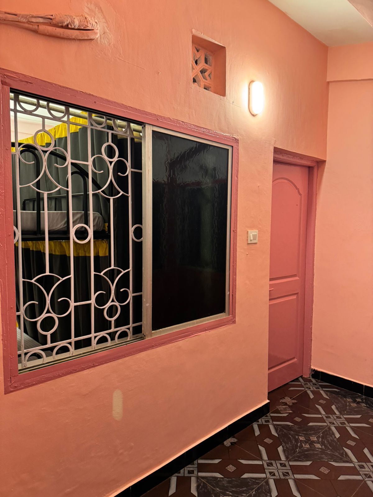 A corridor with a pink wall, a window with decorative metal bars, a door, and patterned floor tiles.