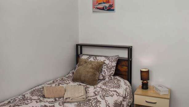 A single bed with patterned bedding, a pillow, two towels, a nightstand with a lamp, and a wall-mounted picture of a car and palm tree.