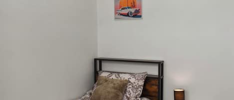 Una cama individual con ropa de cama estampada, una almohada, dos toallas, una mesita de noche con lámpara, y un cuadro en la pared de un coche y una palmera.